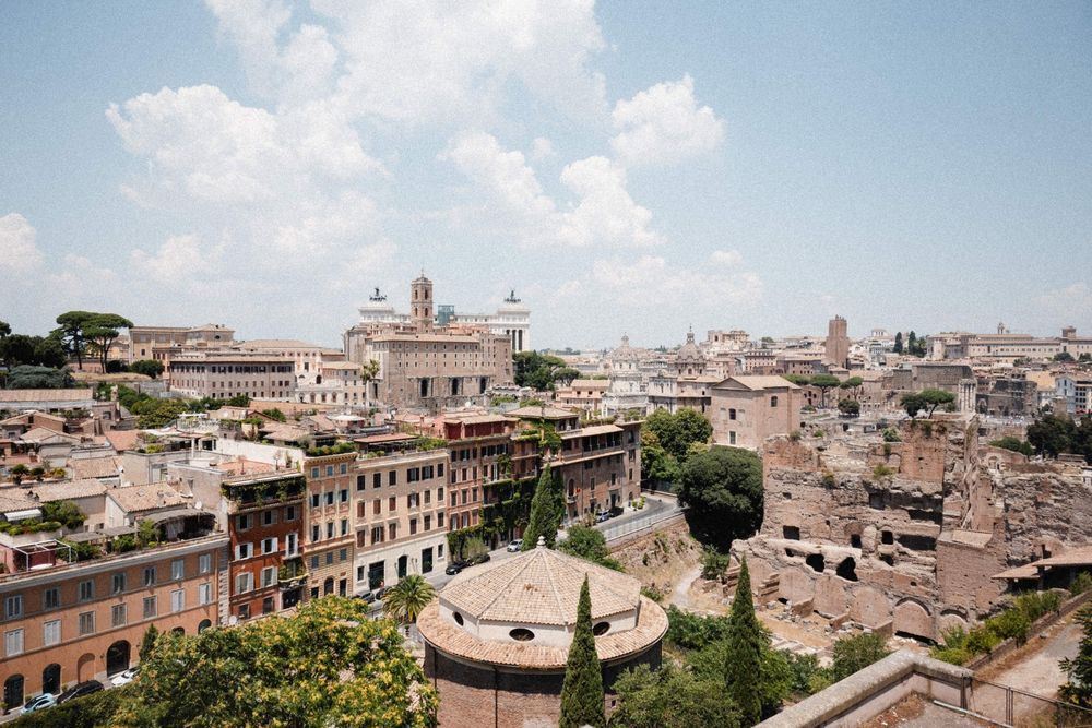 Panoramic view of Rome's historic skyline with ancient buildings, domes, and lush trees under a partly cloudy sky.