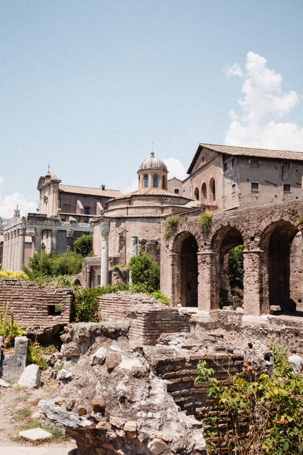 Ancient Roman Forum ruins with arched stone structures, a domed church, and lush greenery under a bright blue sky.
