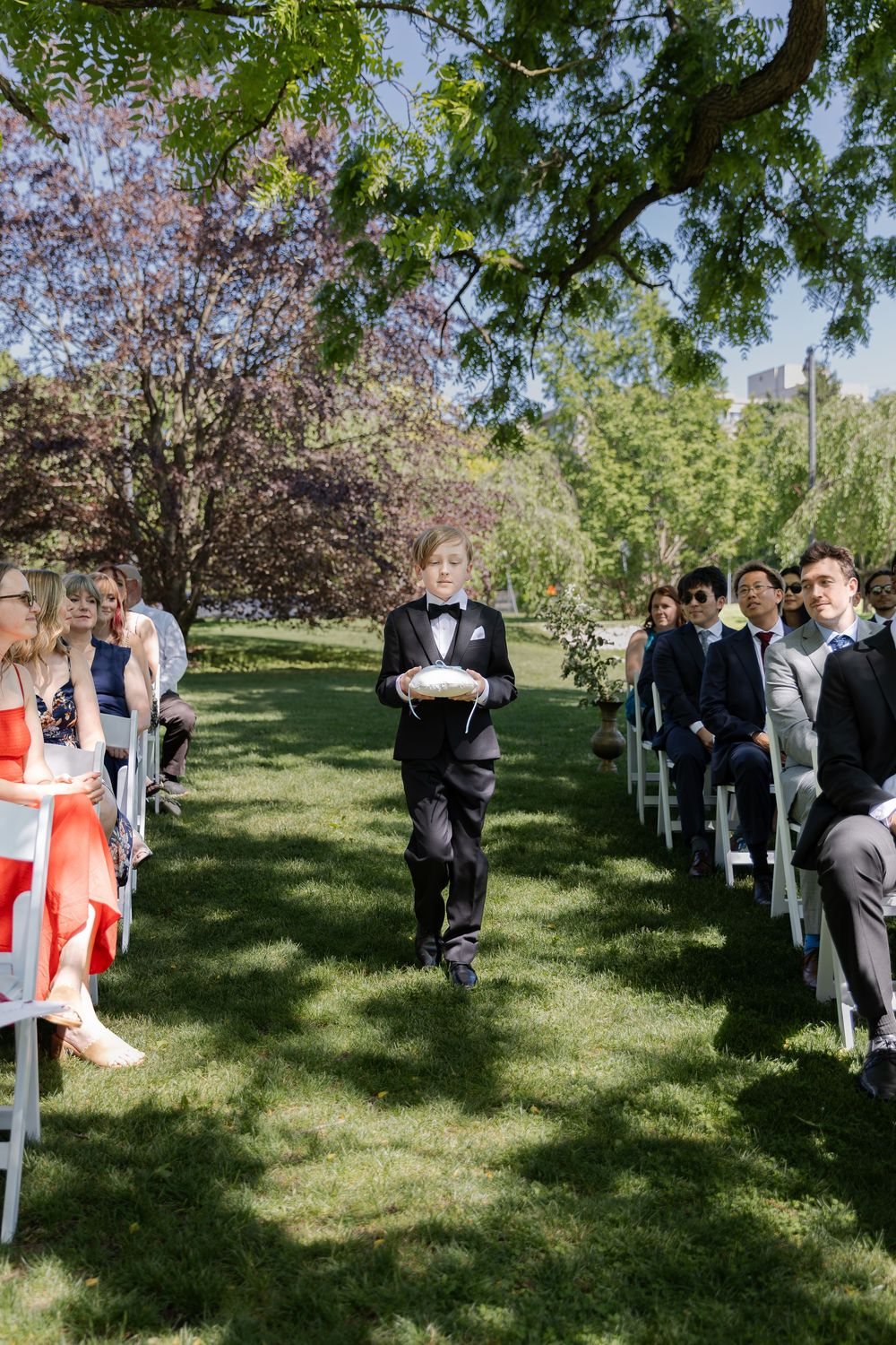 Ring bearer walking down aisle at an outdoor ceremony under the trees at the civic garden complex in london, ontario.