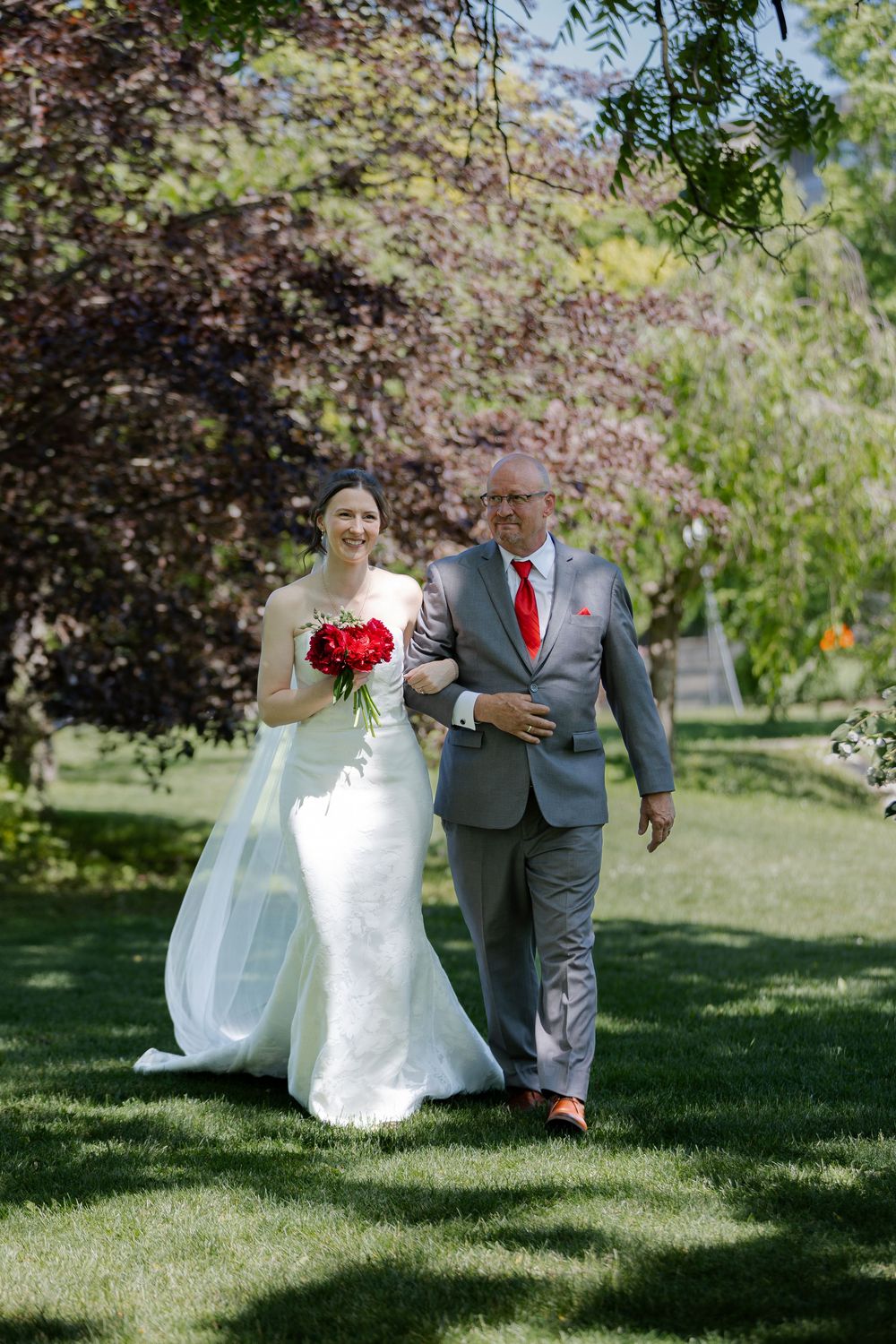 Brides father walks her down the aisle at an outdoor wedding ceremony under the trees at the civic garden complex in london, ontario.