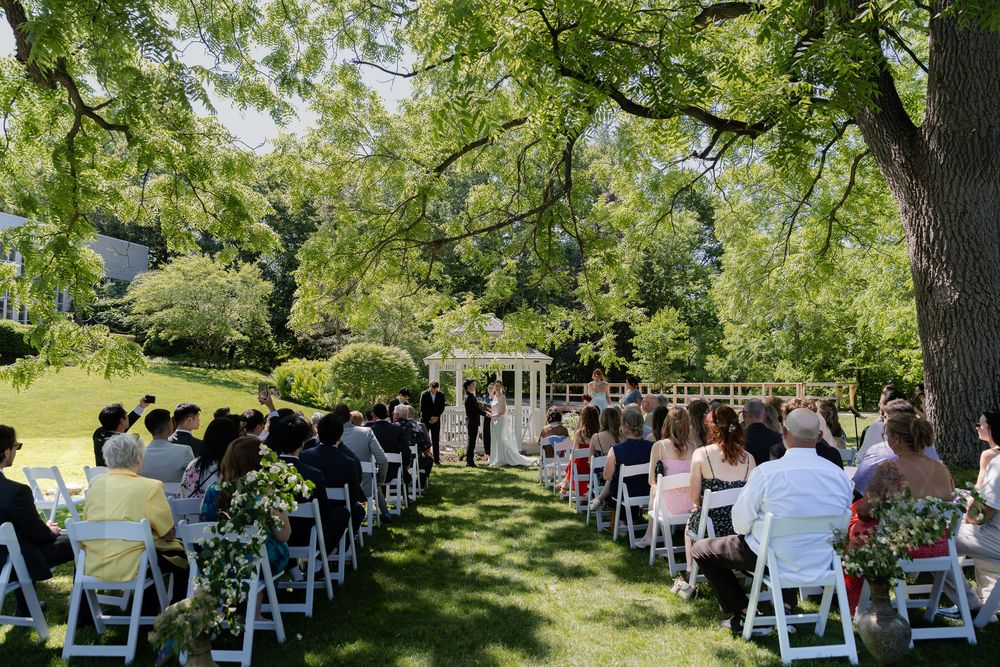 Outdoor wedding ceremony with white chairs, large shade tree, and gazebo surrounded by guests at the civic garden complex in london, ontario