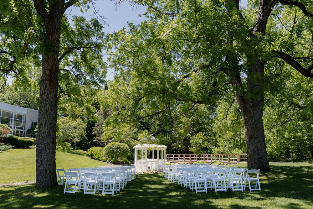 Outdoor wedding ceremony with white chairs on lush green lawn between two large trees with a white gazebo backdrop at civic garden complex.