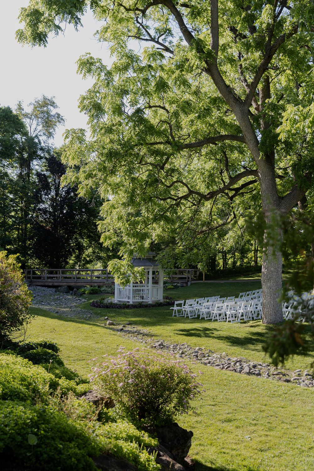 Lush garden wedding venue with white chairs, gazebo, stone pathway, and tall trees on a sunny day at civic garden complex in london ontario.