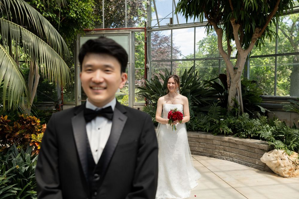 Smiling groom standing with bride in the background during first look in the conservatory at the civic garden complex in london, ontario.