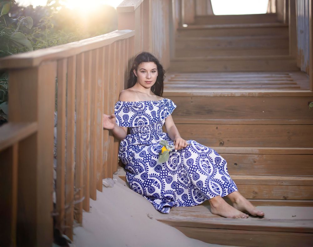 Young woman in blue and white off-shoulder dress sitting gracefully on wooden stairs in golden sunlight.