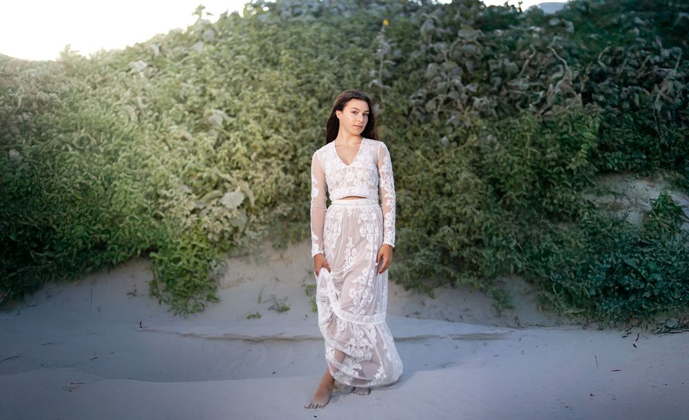 Woman in white lace gown standing on sandy beach dunes surrounded by lush green coastal shrubs.