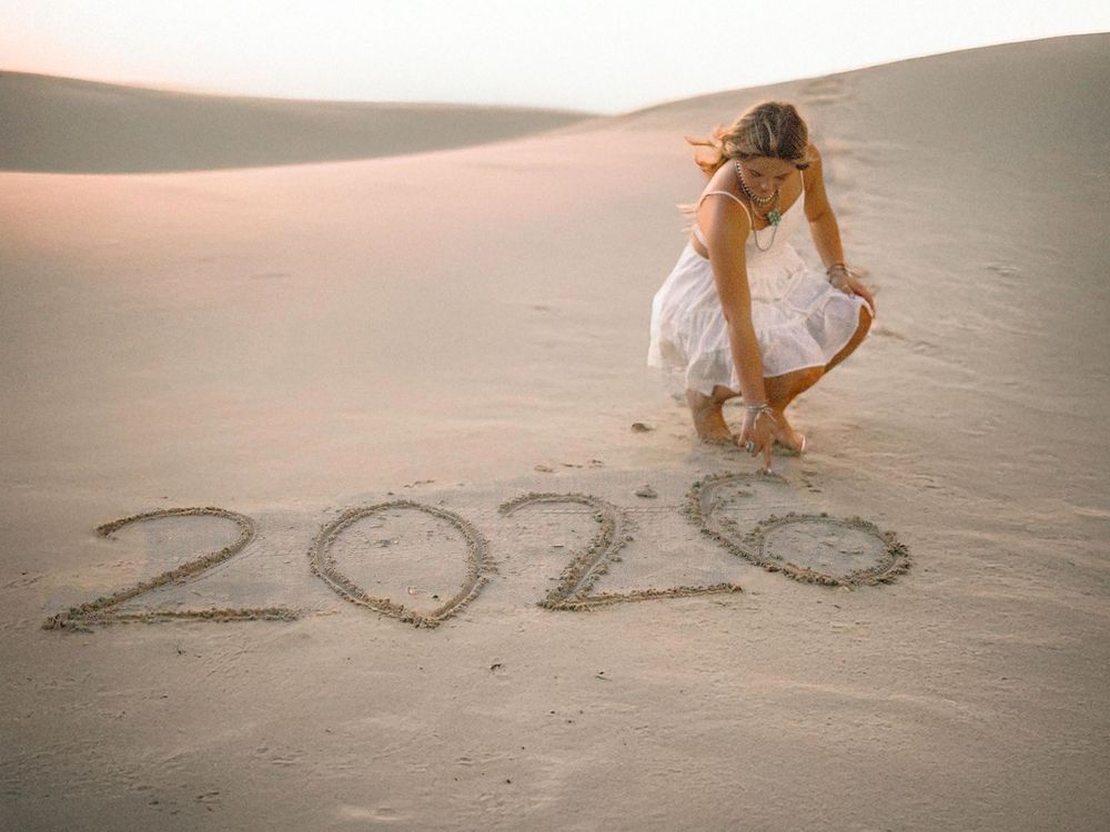 Woman in white dress writing 2026 in sand on desert dunes at sunset.