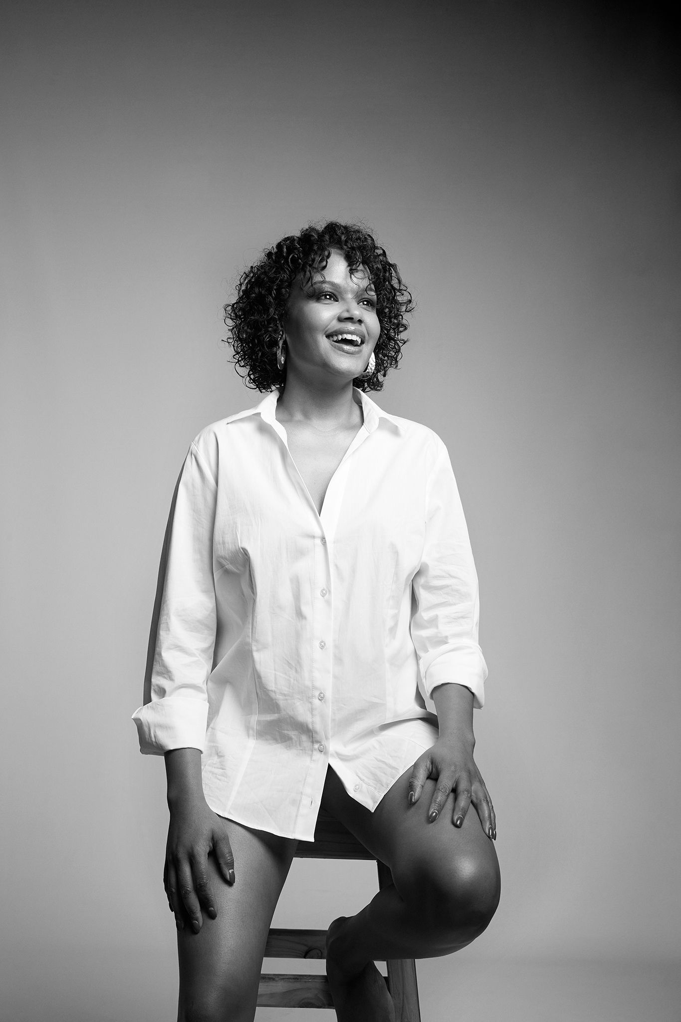 A smiling Black woman with curly hair sits on a stool wearing an oversized white shirt in a black and white portrait.