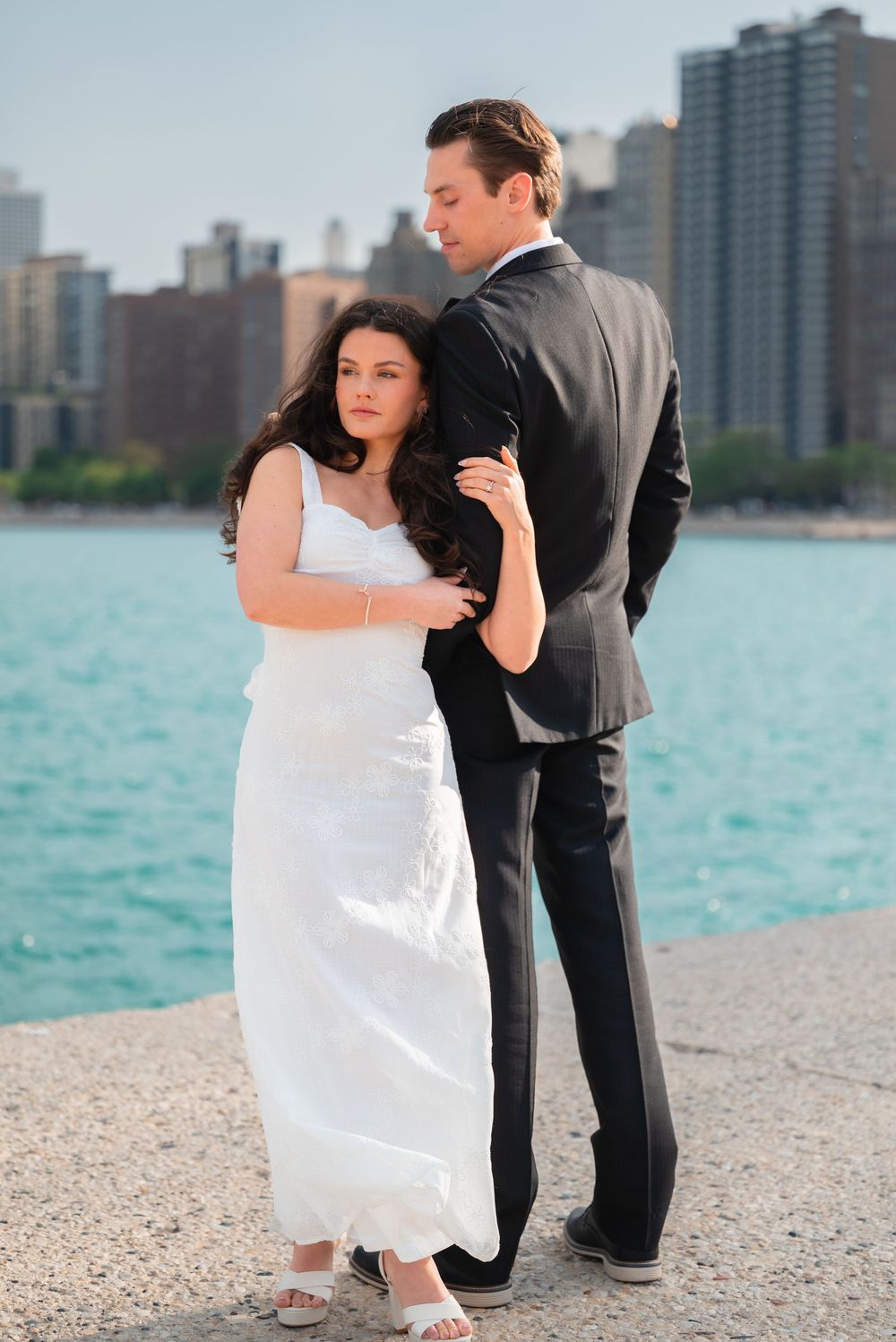Bride in white gown and groom in black suit pose by waterfront with city skyline backdrop.