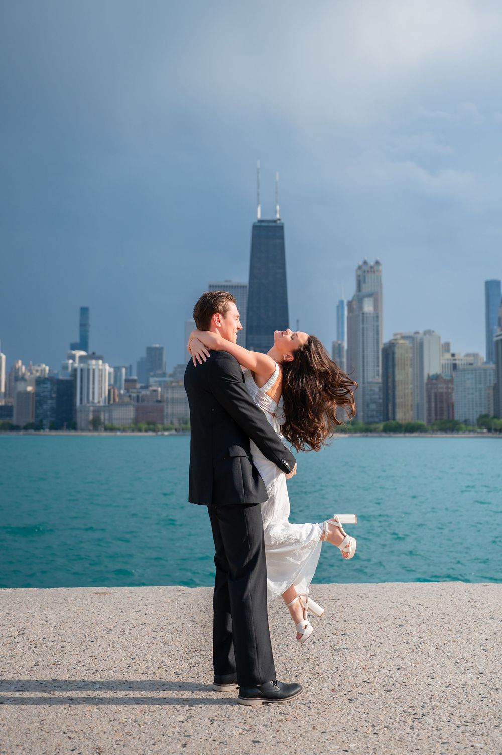 Bride and groom embrace on Chicago lakefront with iconic skyline and John Hancock Center in background.