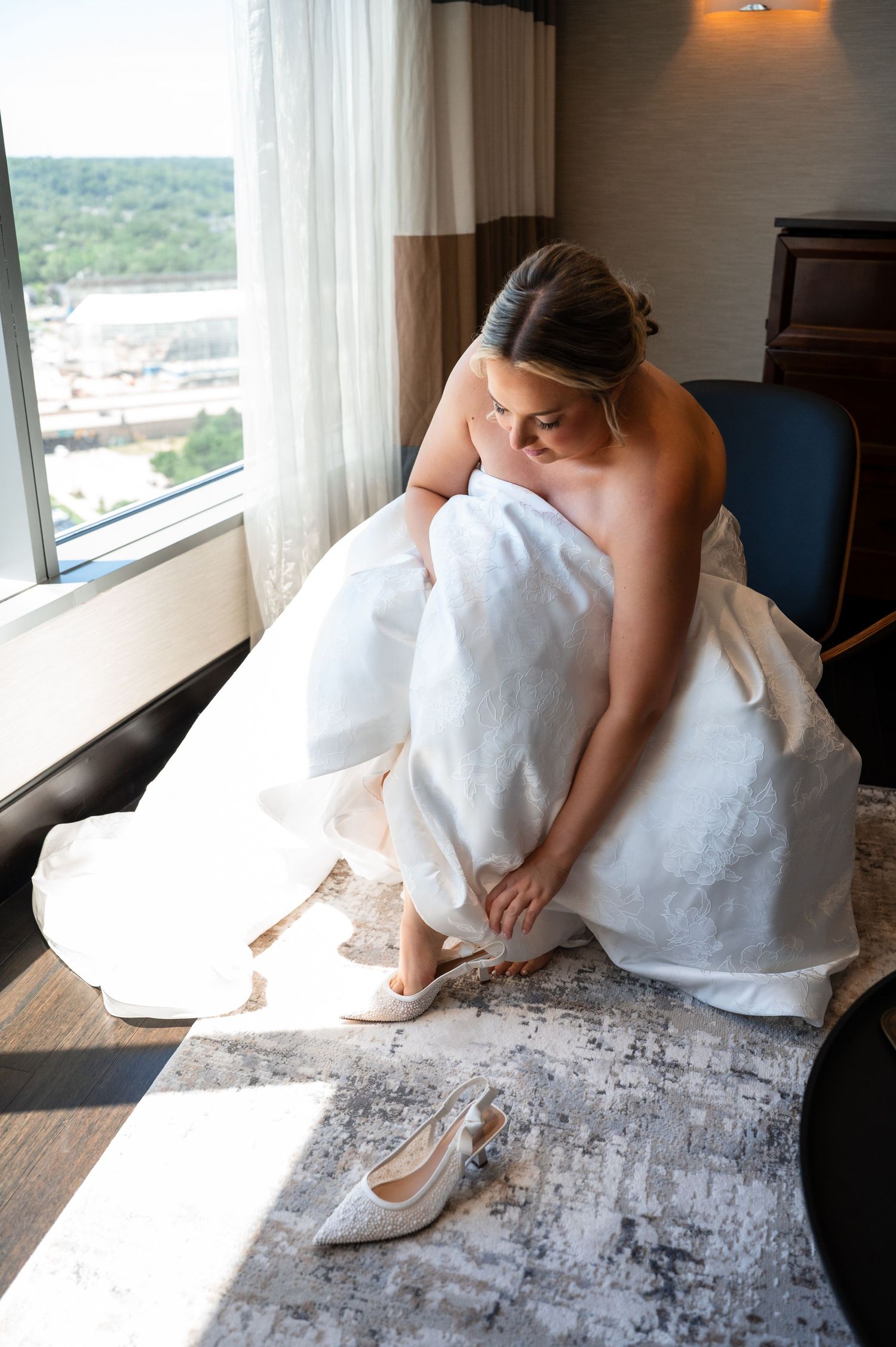 Bride in white gown putting on heels by hotel window on wedding day.