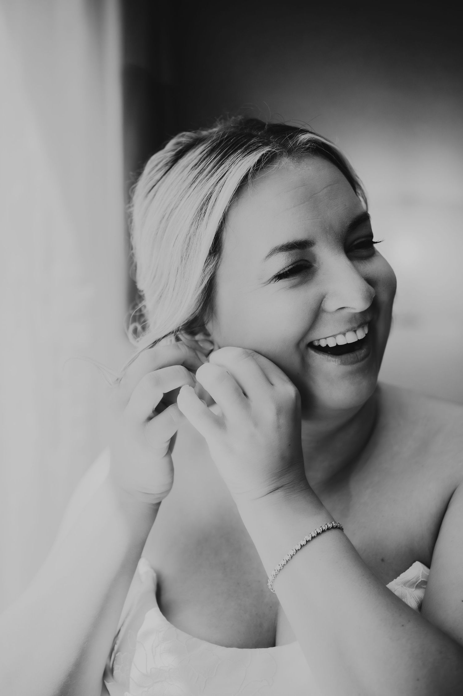 Black and white portrait of laughing blonde bride putting on earrings while getting ready for wedding.