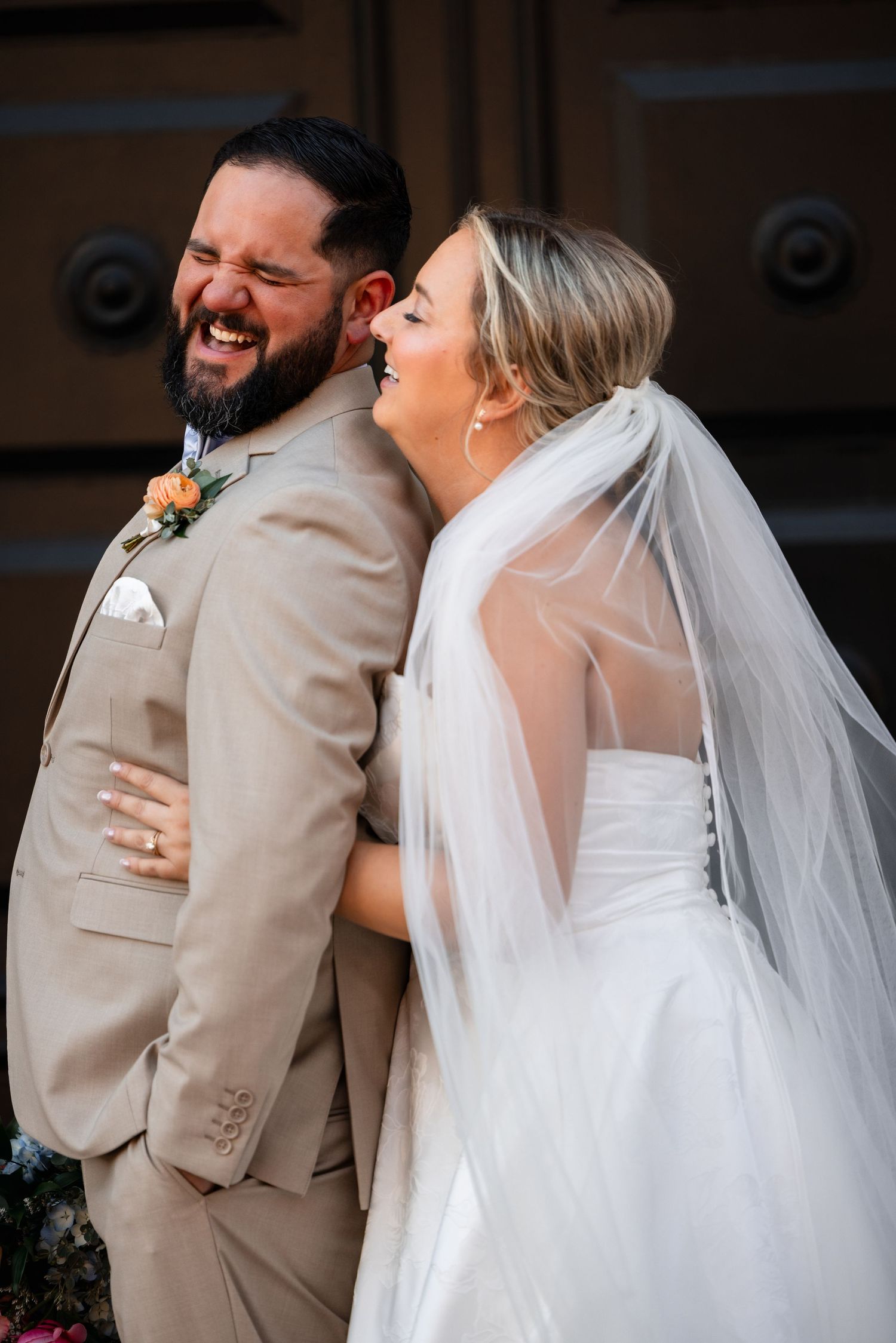 Laughing bride and groom embrace outside wooden doors, groom in tan suit with floral boutonniere, bride in white gown with veil.