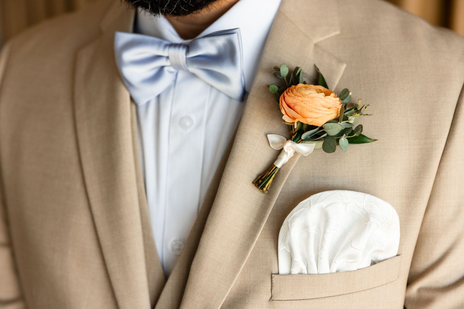 Close-up of groom in tan suit with blue bow tie, orange ranunculus boutonniere, and white pocket square.