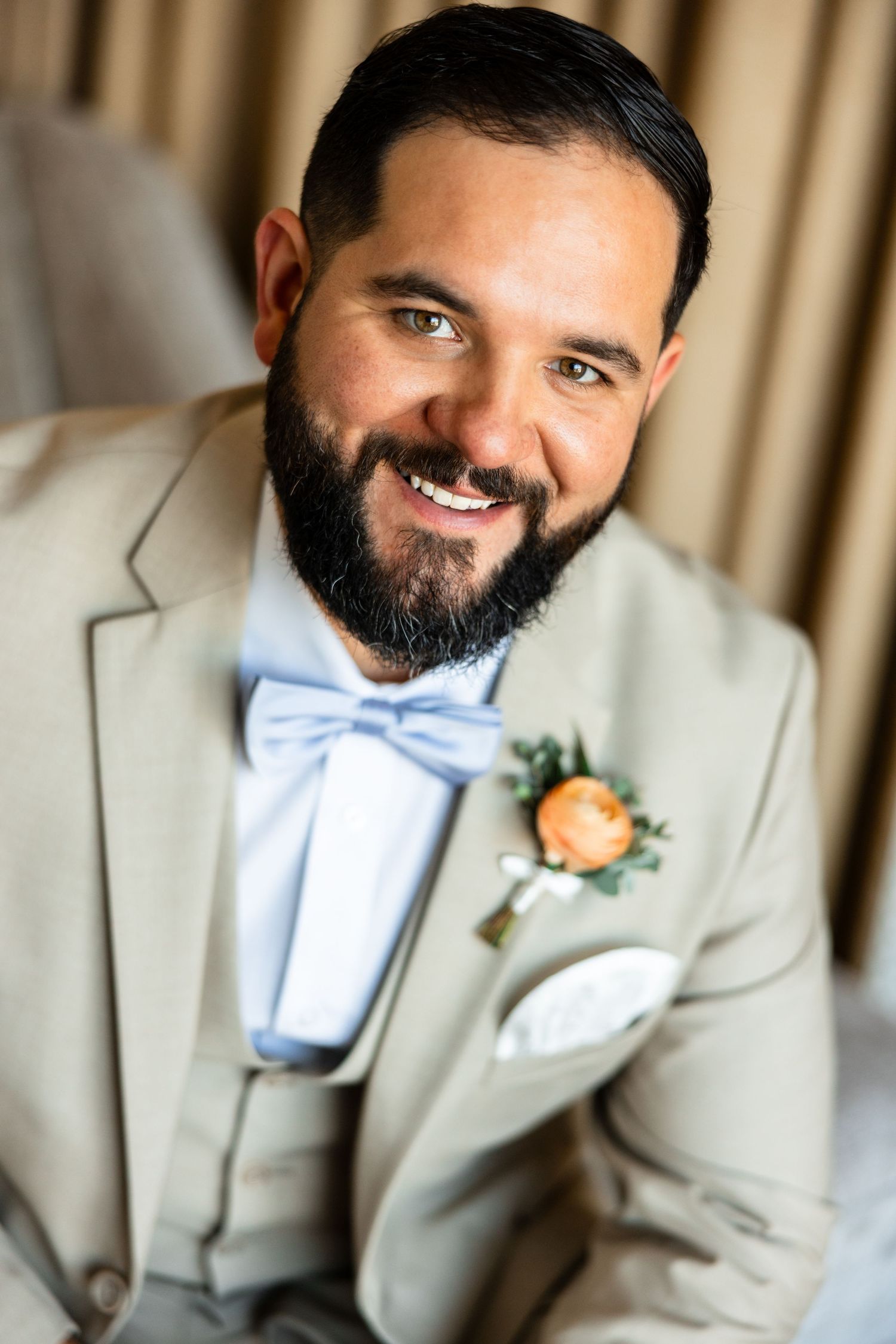 Smiling bearded groom in tan suit with blue bow tie and orange boutonniere poses for wedding portrait.