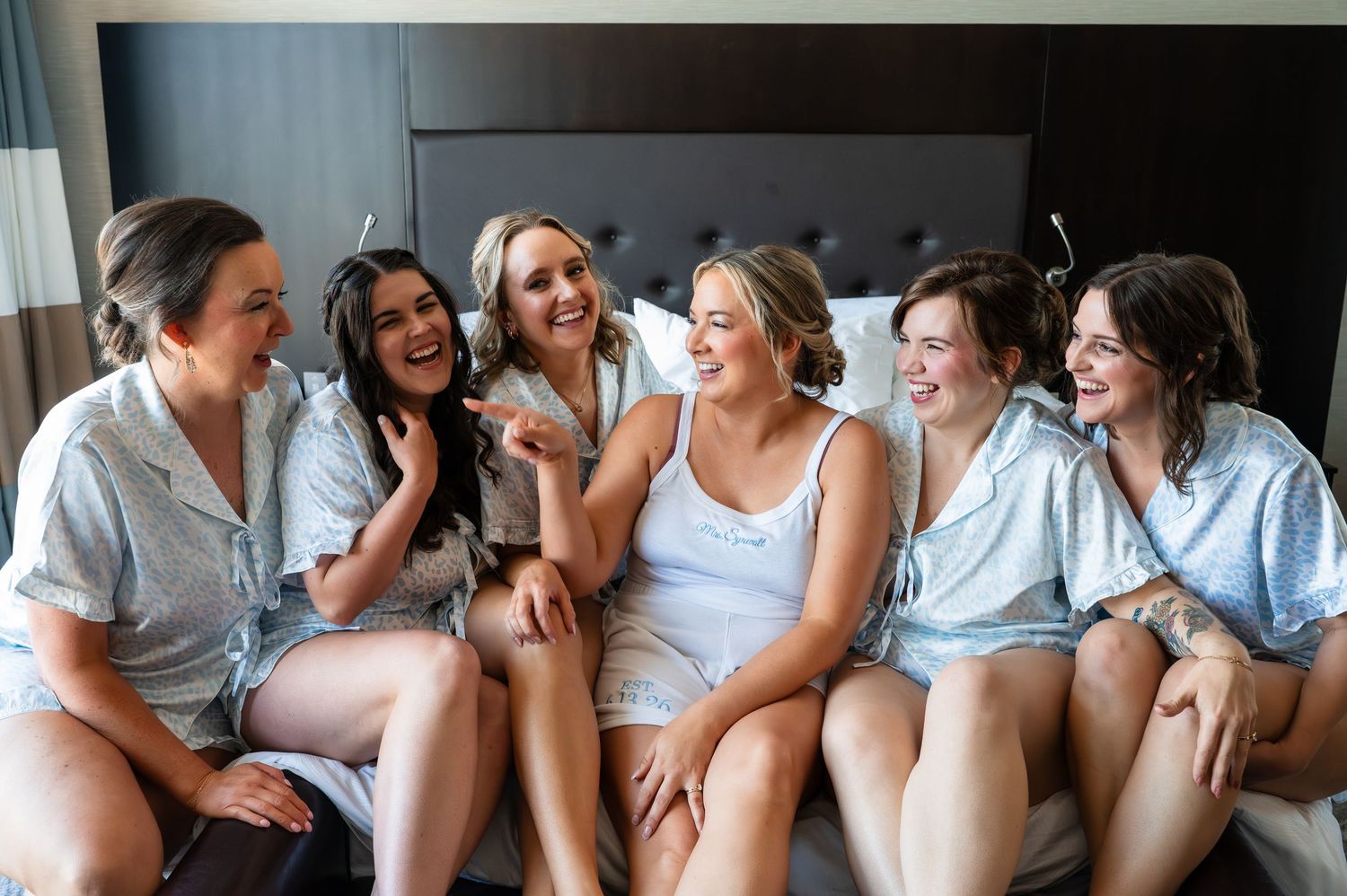 Five bridesmaids in matching pajamas laughing together on a hotel bed during a bridal party getting-ready session.