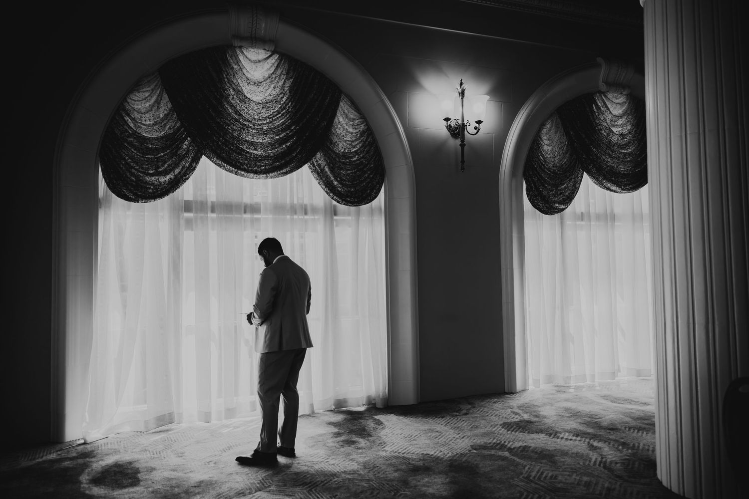 Black and white photo of a man standing alone by large arched windows with draped curtains in an elegant room.