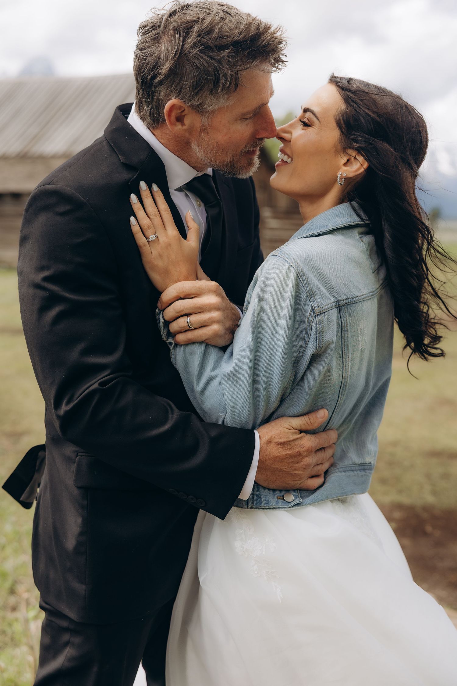 A bride in a denim jacket and white skirt embraces her groom in a black suit outdoors.