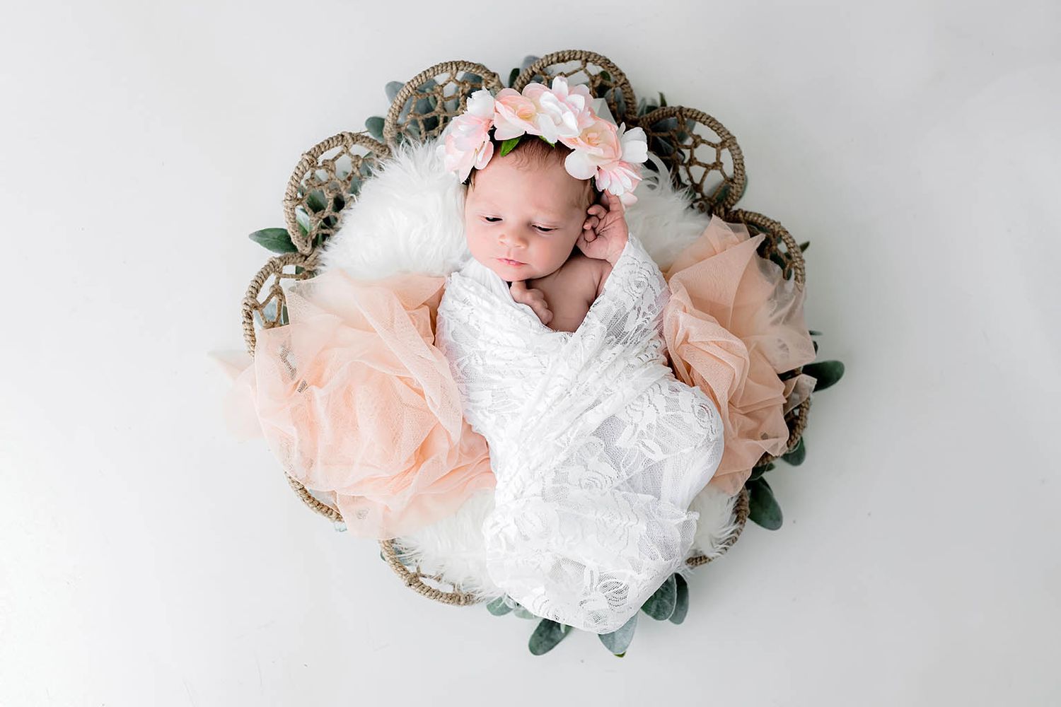 Newborn baby girl sleeping in a wicker basket with pink floral headband, white lace wrap, and soft peach tulle.