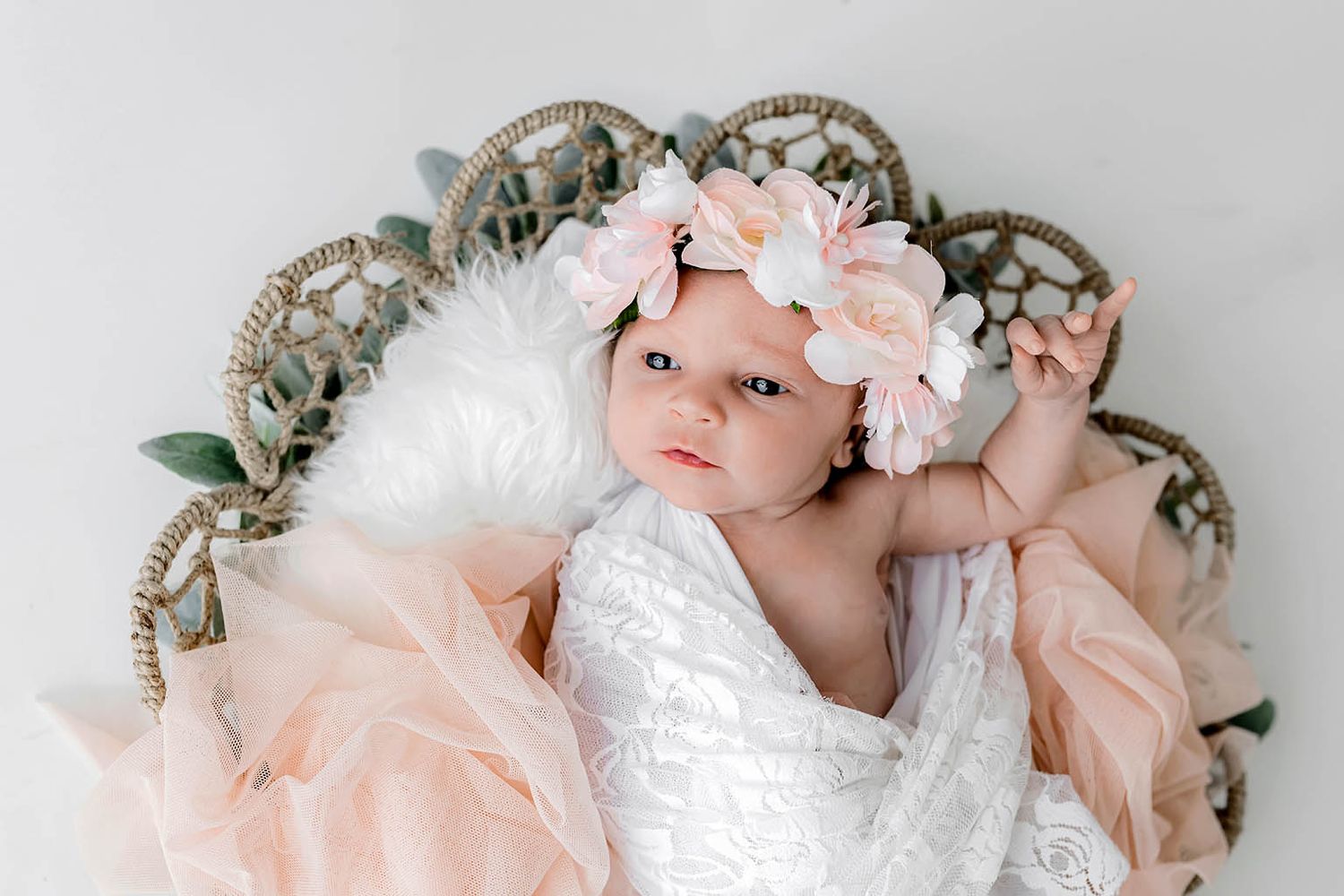 Newborn baby girl in floral headband wrapped in white lace, resting in a wicker basket with blush pink tulle.
