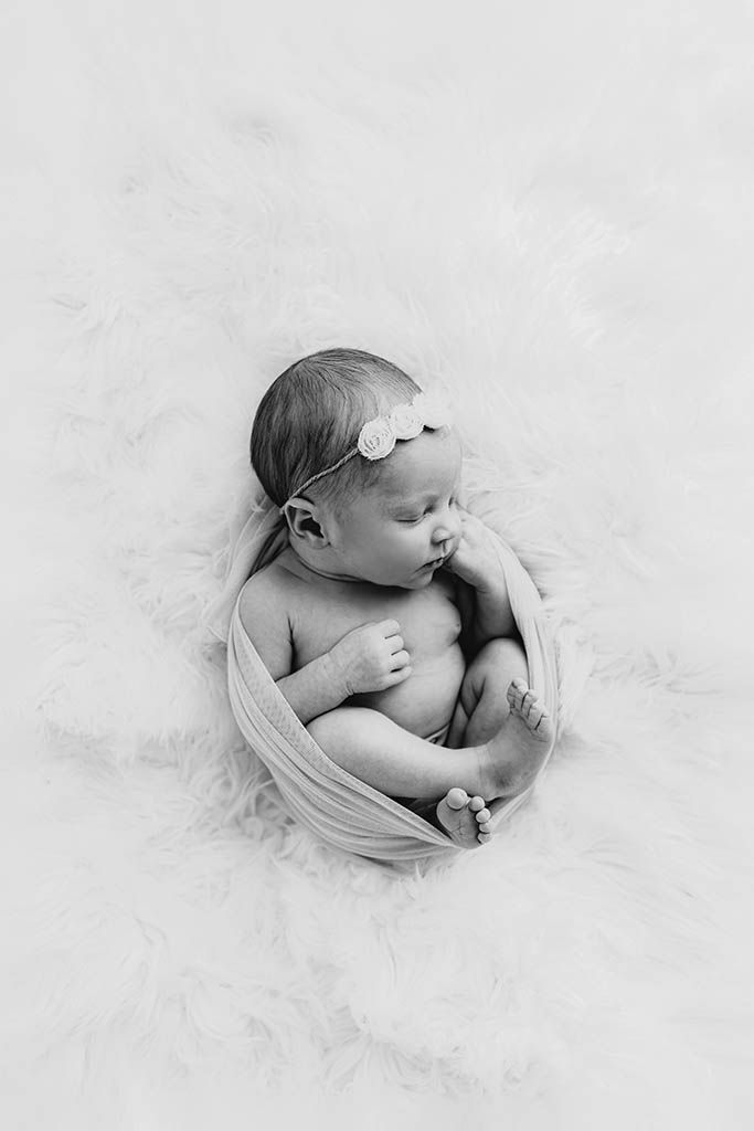 Black and white newborn photo of a baby girl wrapped in soft fabric with a floral headband, resting on a fluffy white rug.