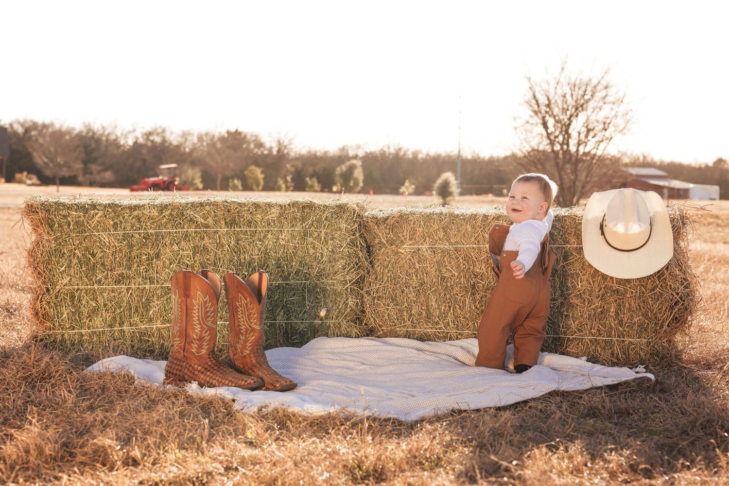 Baby boy in suspenders sits on blanket by hay bales with cowboy boots and hat in golden field.