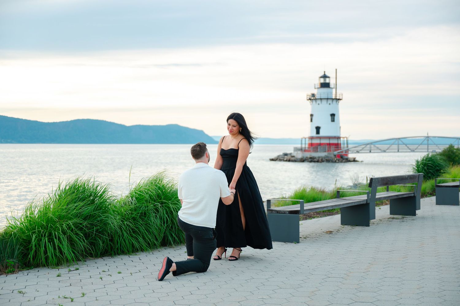 Man proposing on one knee to woman in black dress by a lighthouse on the Hudson River waterfront.