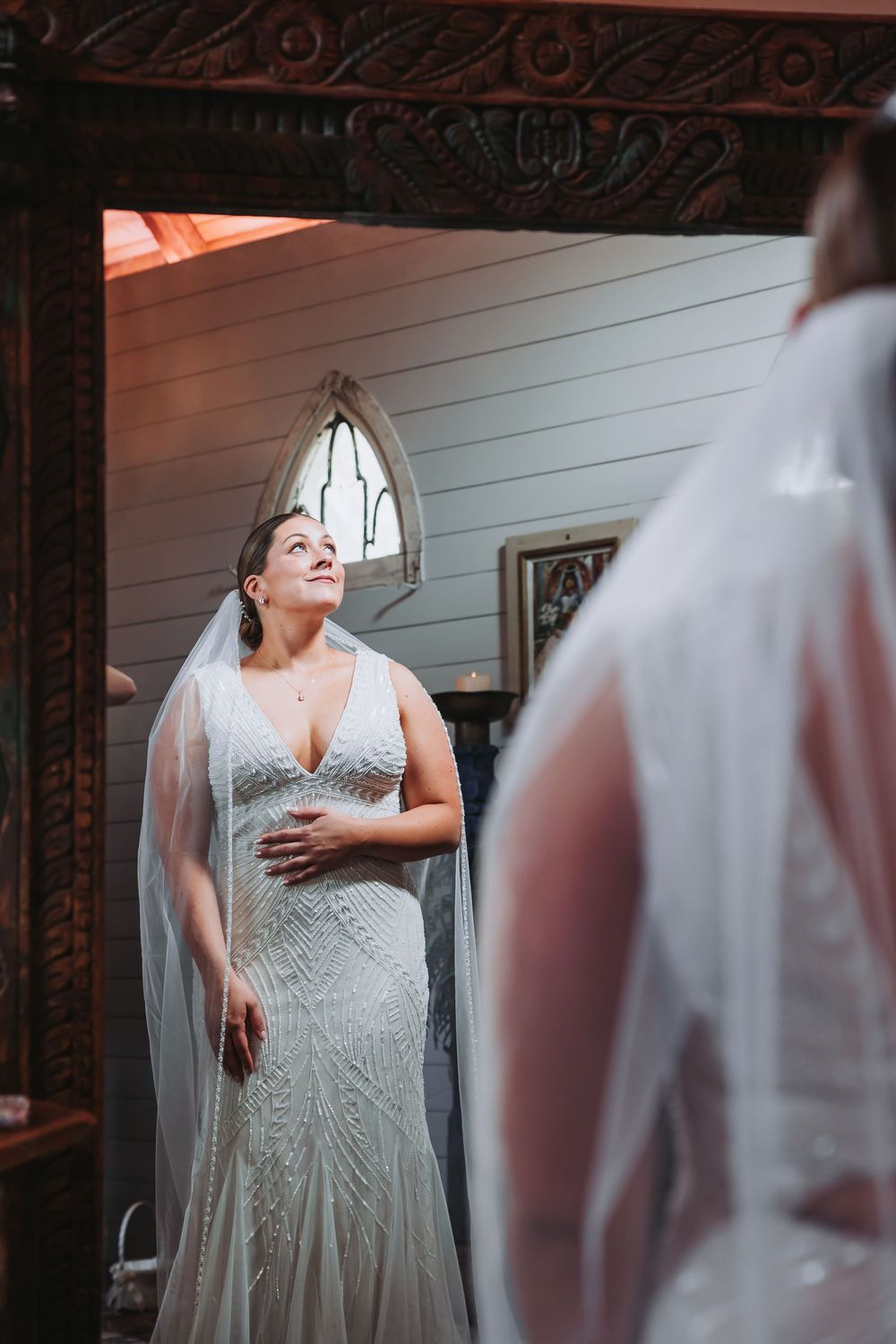 Bride in beaded gown and veil gazes upward while viewing her reflection in an ornate mirror before her wedding.