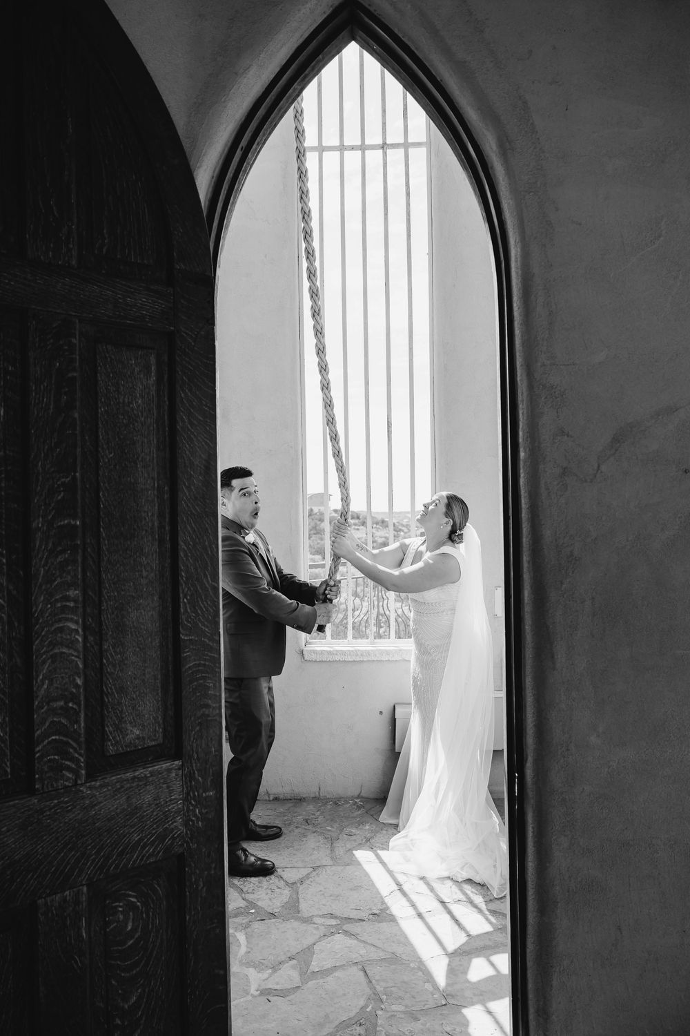 Black and white photo of bride and groom holding hands framed by gothic arch doorway at wedding venue.