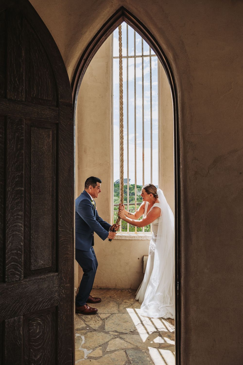 Bride and groom hold hands through a Gothic arch doorway in a sunlit chapel during their wedding ceremony.