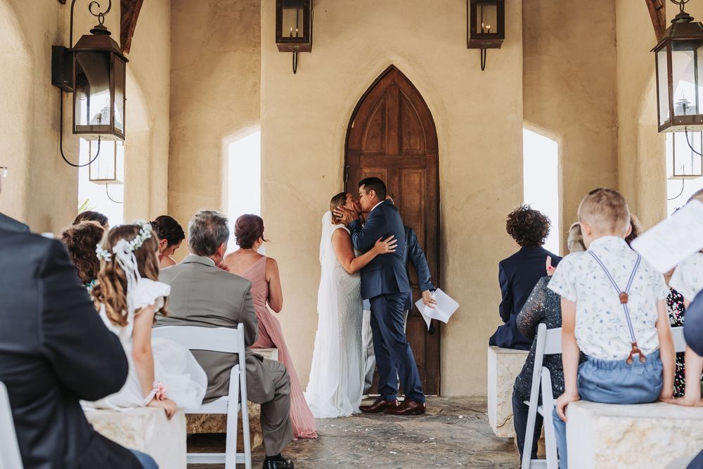 Bride and groom share first kiss at rustic chapel altar surrounded by seated wedding guests.