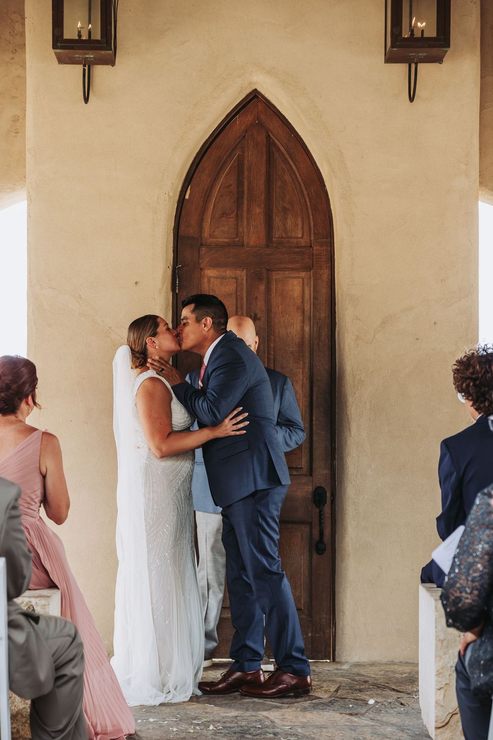Bride and groom share their first kiss at the altar in front of a wooden arched door, surrounded by wedding guests.