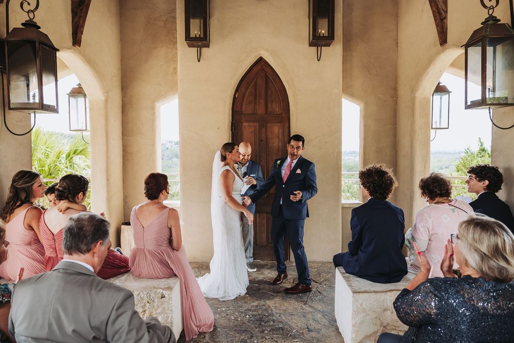 Bride and groom exchange vows at an outdoor chapel ceremony with bridesmaids in pink dresses and guests seated nearby.