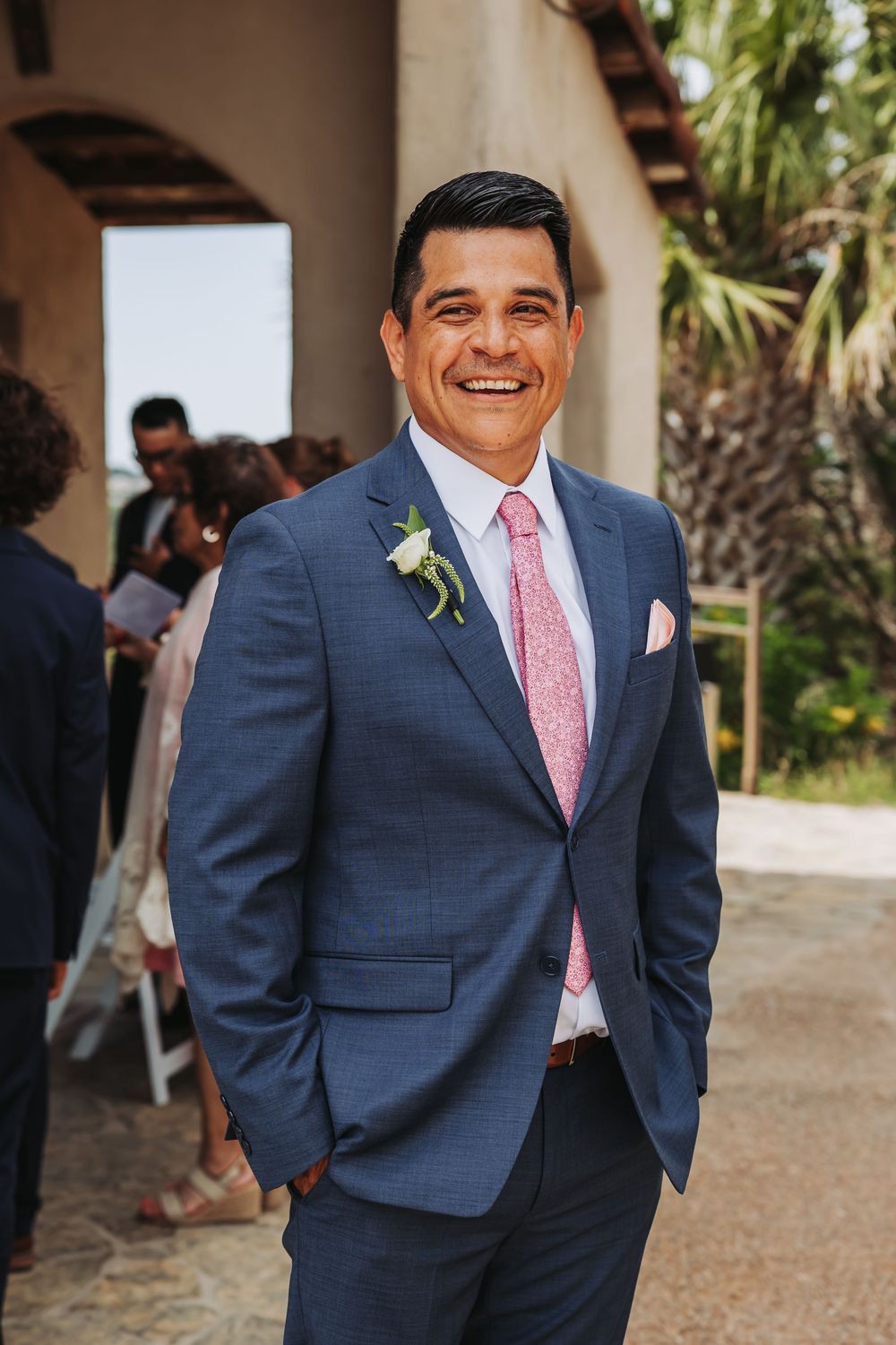 Smiling man in navy blue suit with pink tie and boutonniere at outdoor wedding ceremony.