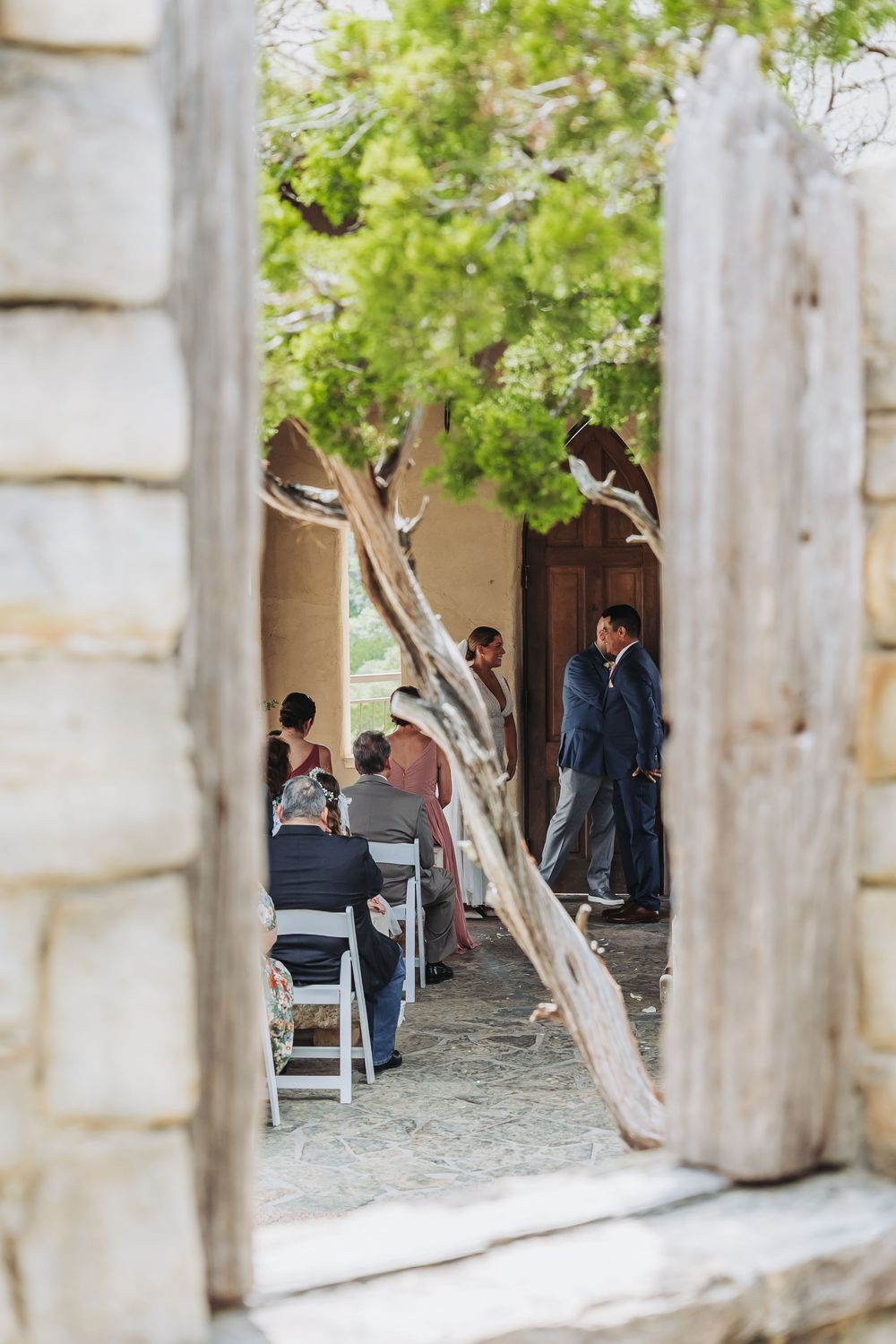 Wedding guests seated outdoors viewed through rustic stone doorway framing.