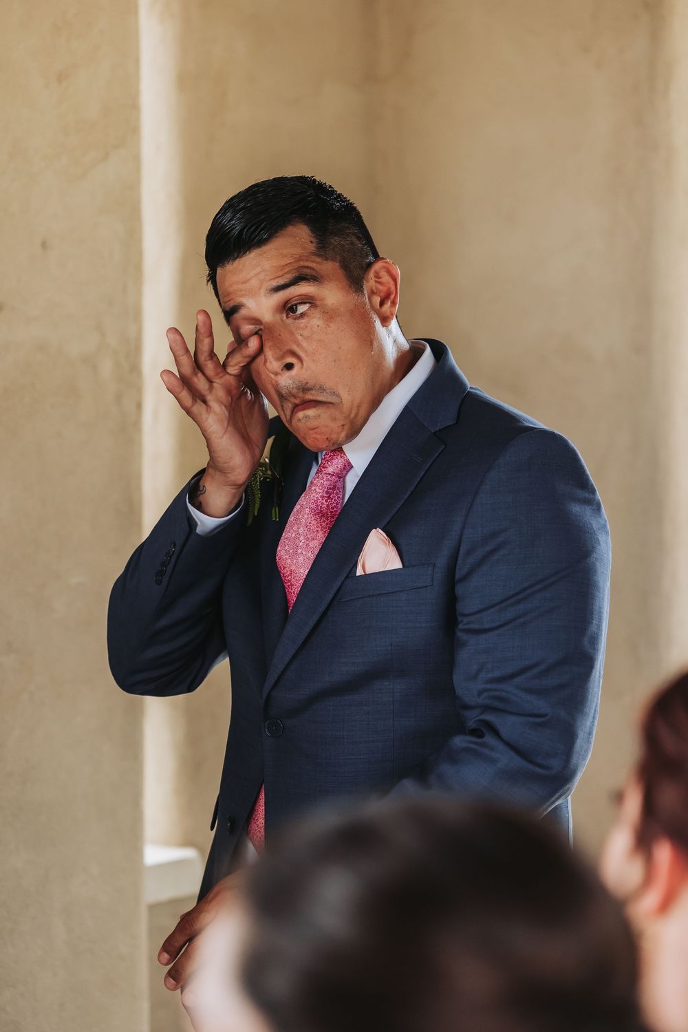 Man in navy suit with pink tie wiping eye at formal event.