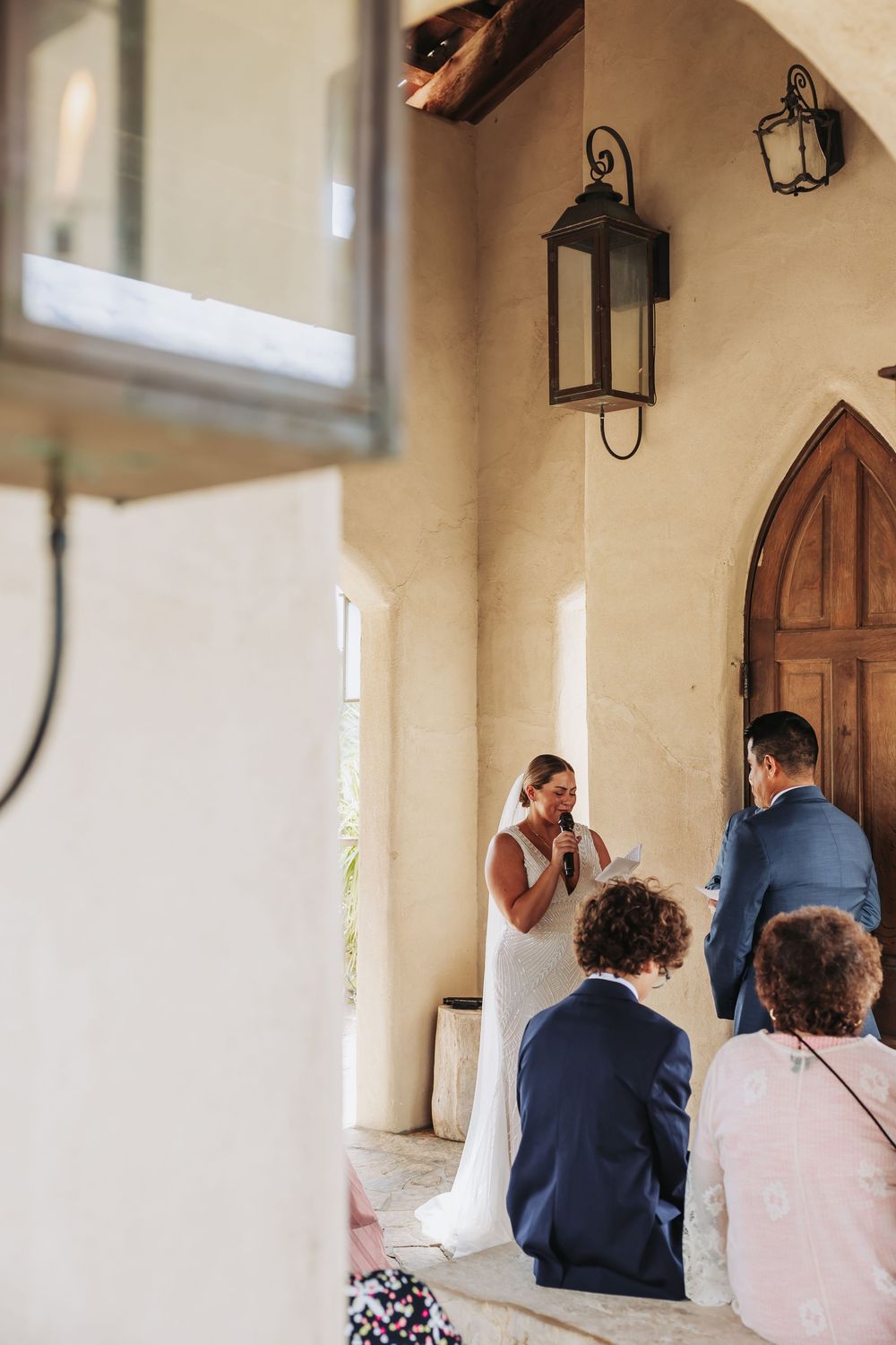 Bride reads vows to groom during outdoor wedding ceremony at rustic chapel with arched wooden doors.