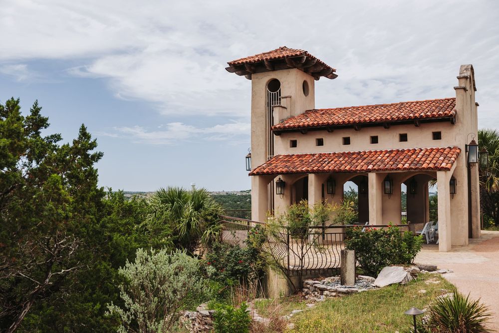 Spanish colonial revival building with red tile roof and bell tower surrounded by lush greenery under cloudy skies.