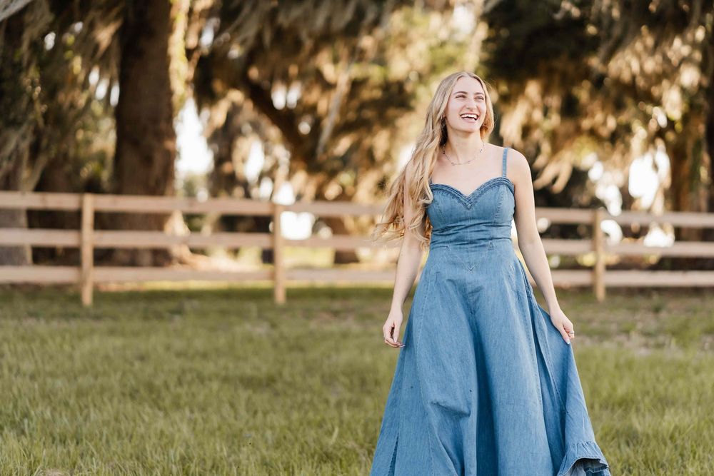 Young woman in elegant blue ball gown smiling outdoors on green grass with wooden fence and trees.