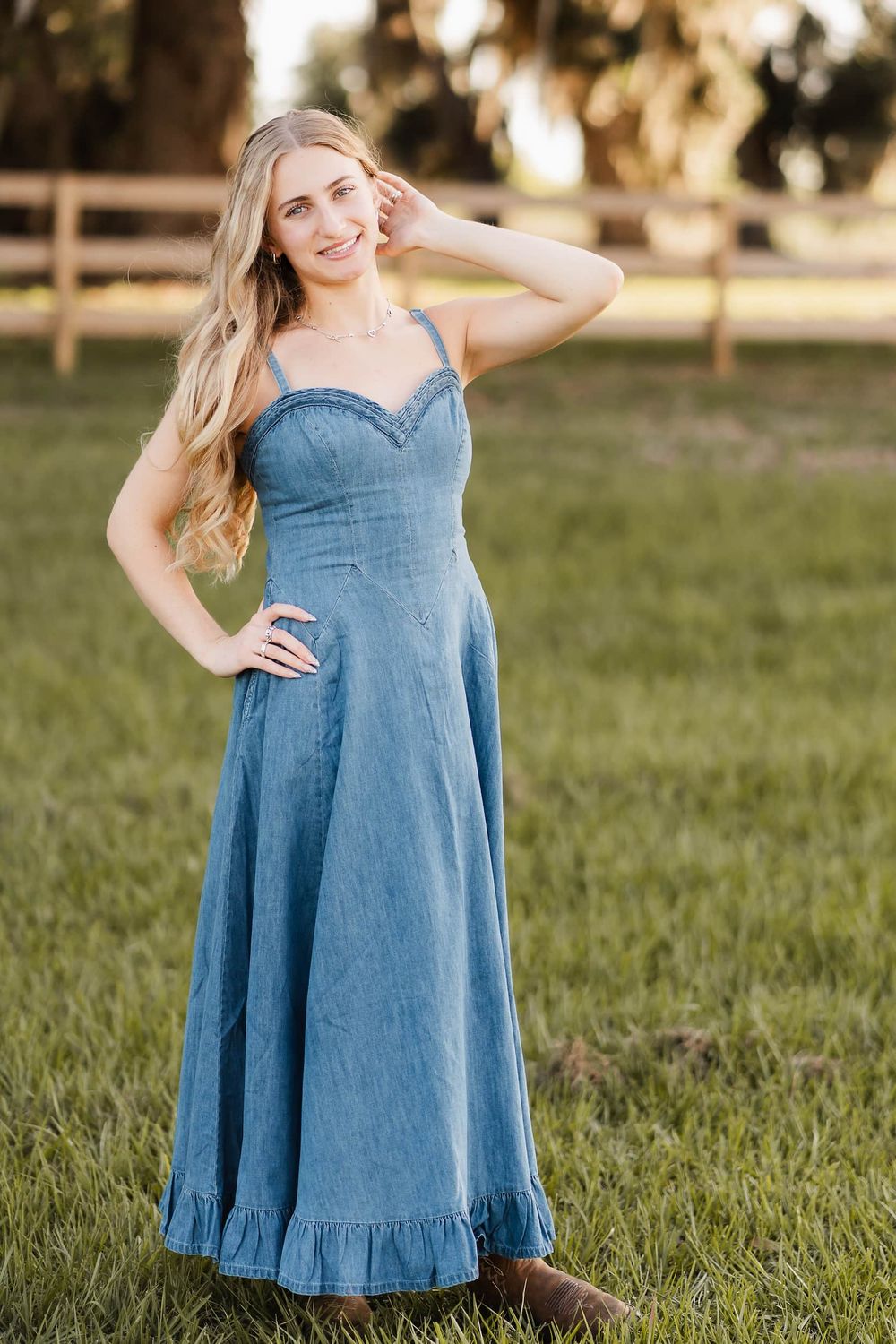 Young woman in denim maxi dress with ruffled hem and cowboy boots posing outdoors on green grass near wooden fence.