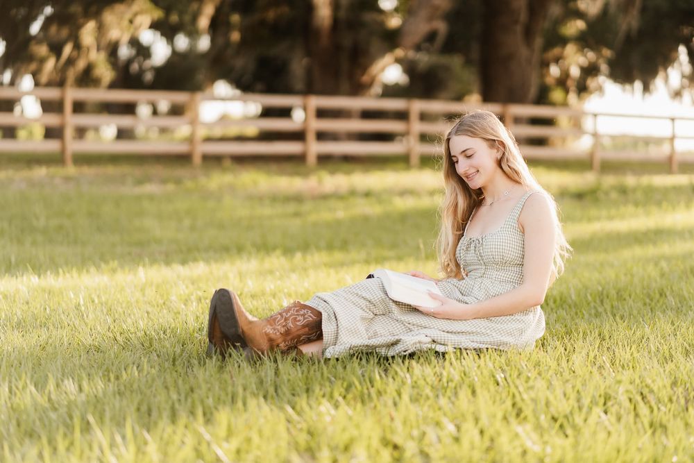 Young woman in a sundress and cowboy boots sitting on lush green grass near a wooden fence on a farm.
