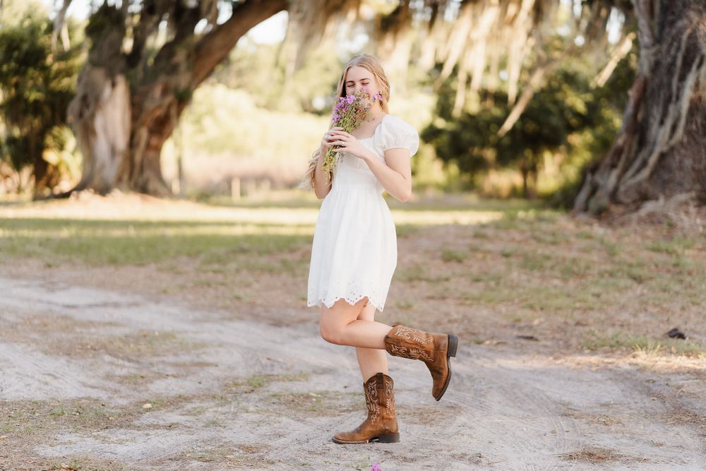 Young blonde woman in white dress and cowboy boots smells wildflowers on a sunlit dirt path under large oak trees.