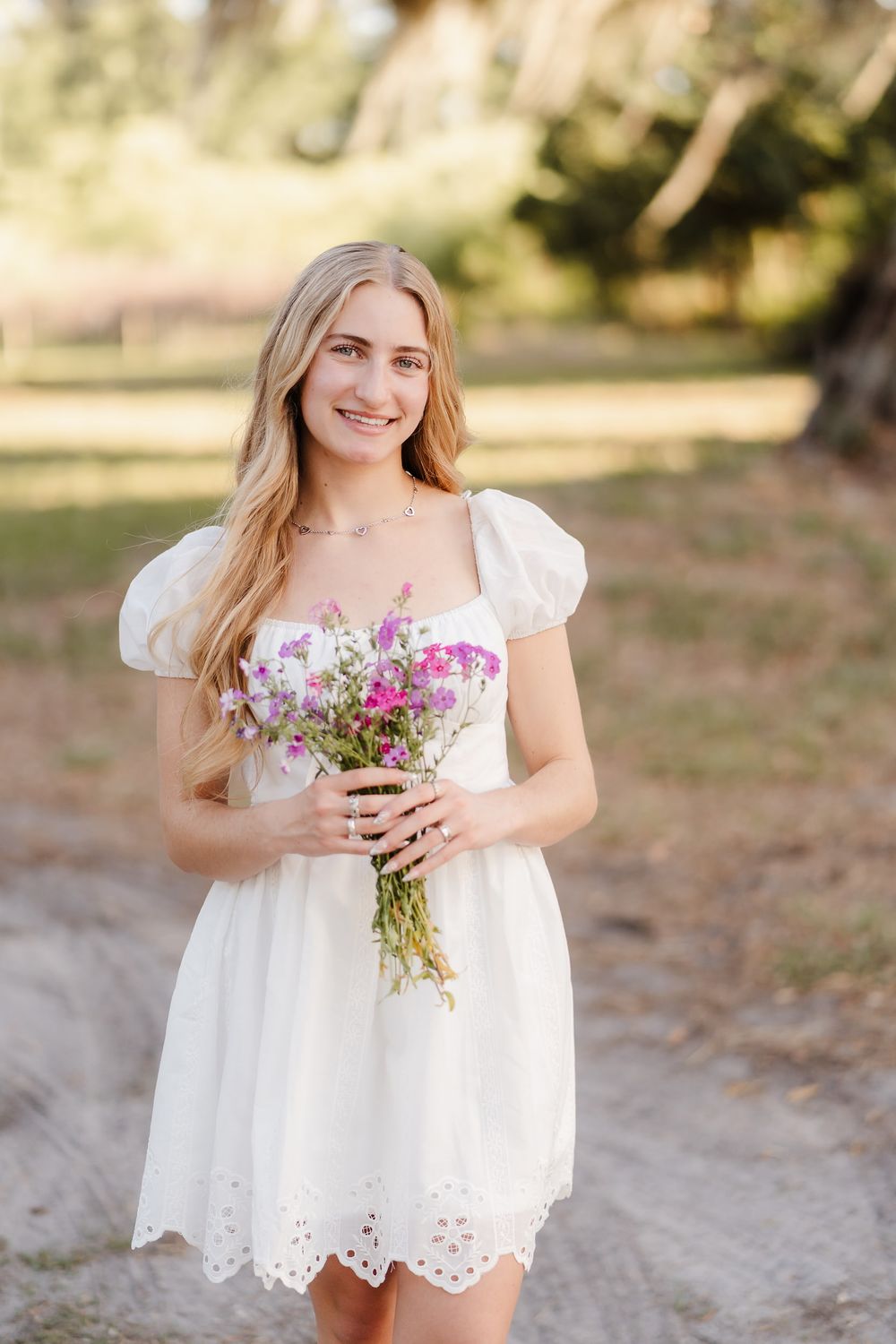 Young blonde woman in white eyelet puff-sleeve mini dress holding wildflower bouquet outdoors on sandy path.
