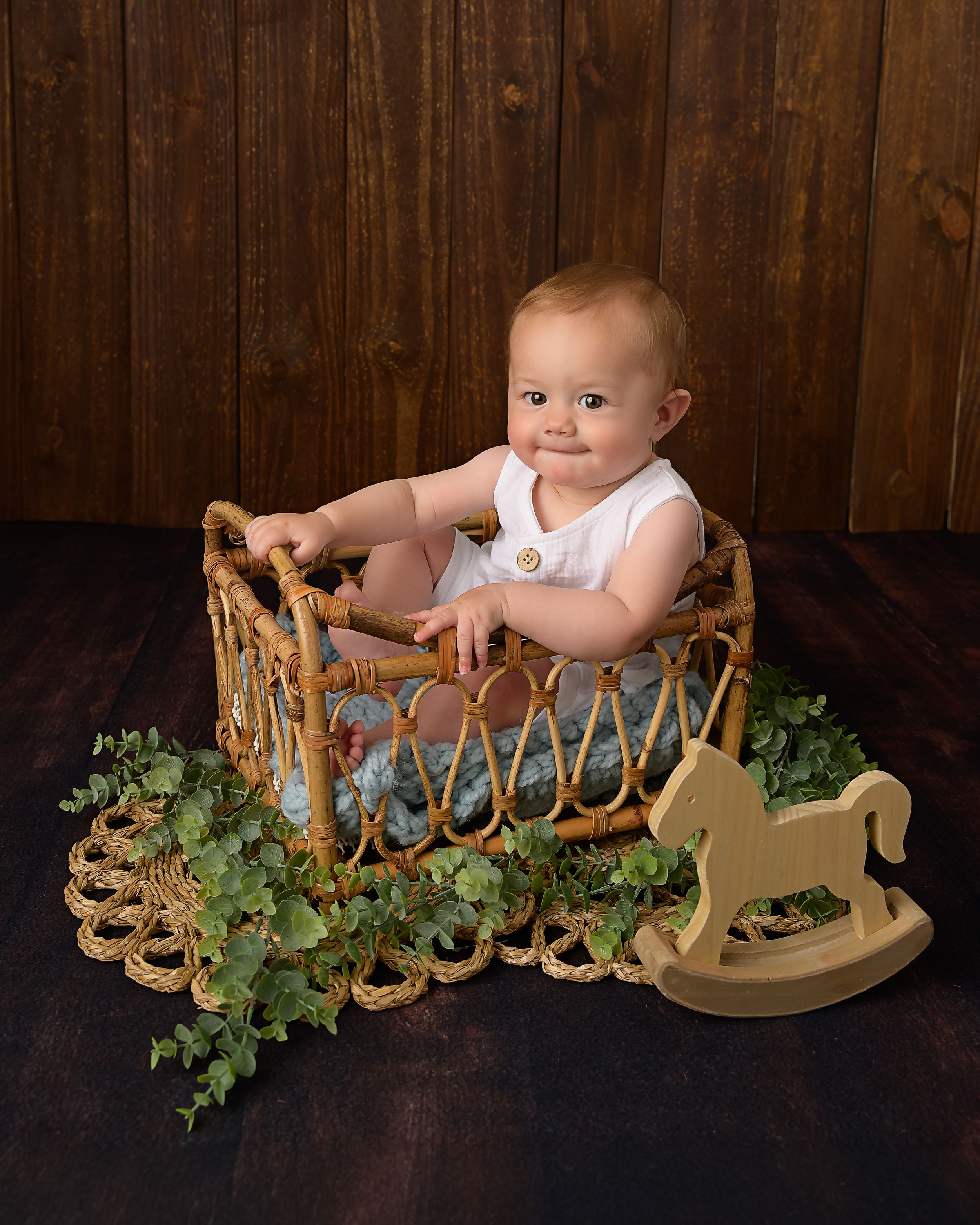 Smiling baby boy sitting in a wicker basket bed with ivy and wooden rocking horse on dark background.