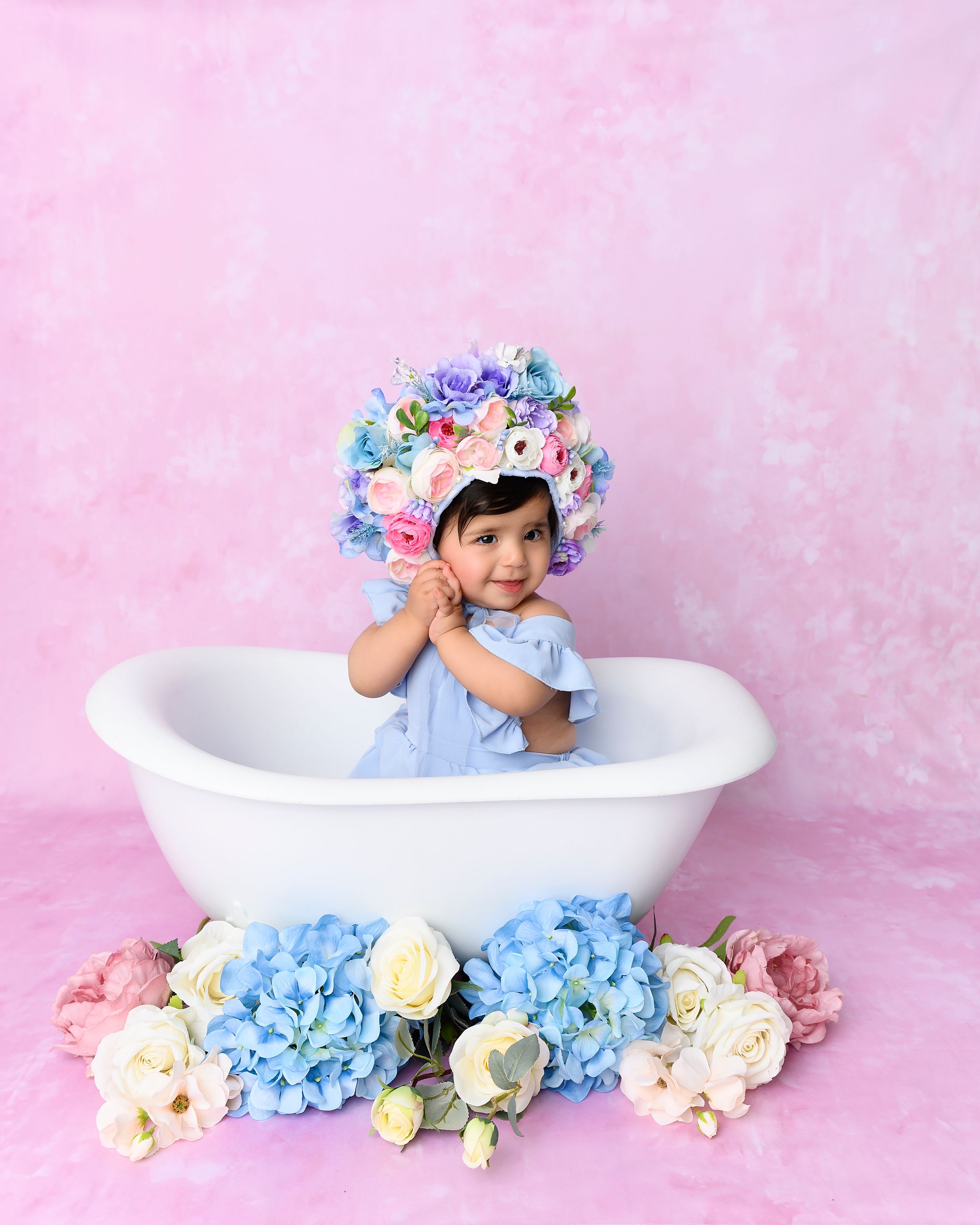 Baby girl in blue dress and floral bonnet sitting in white bathtub with colorful flowers on pink background.