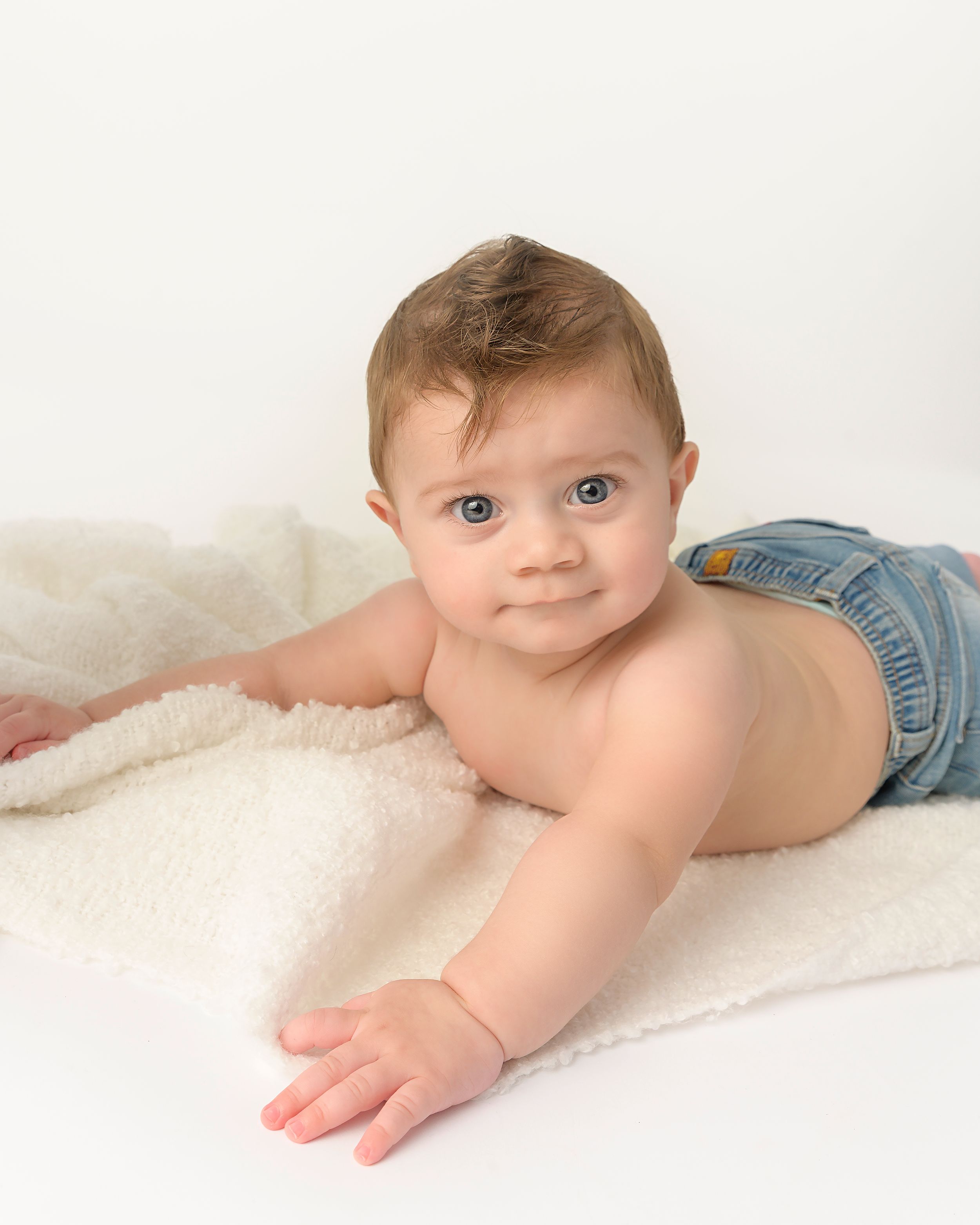 Adorable baby boy with blue eyes lying on fluffy white blanket wearing denim shorts in studio photo.