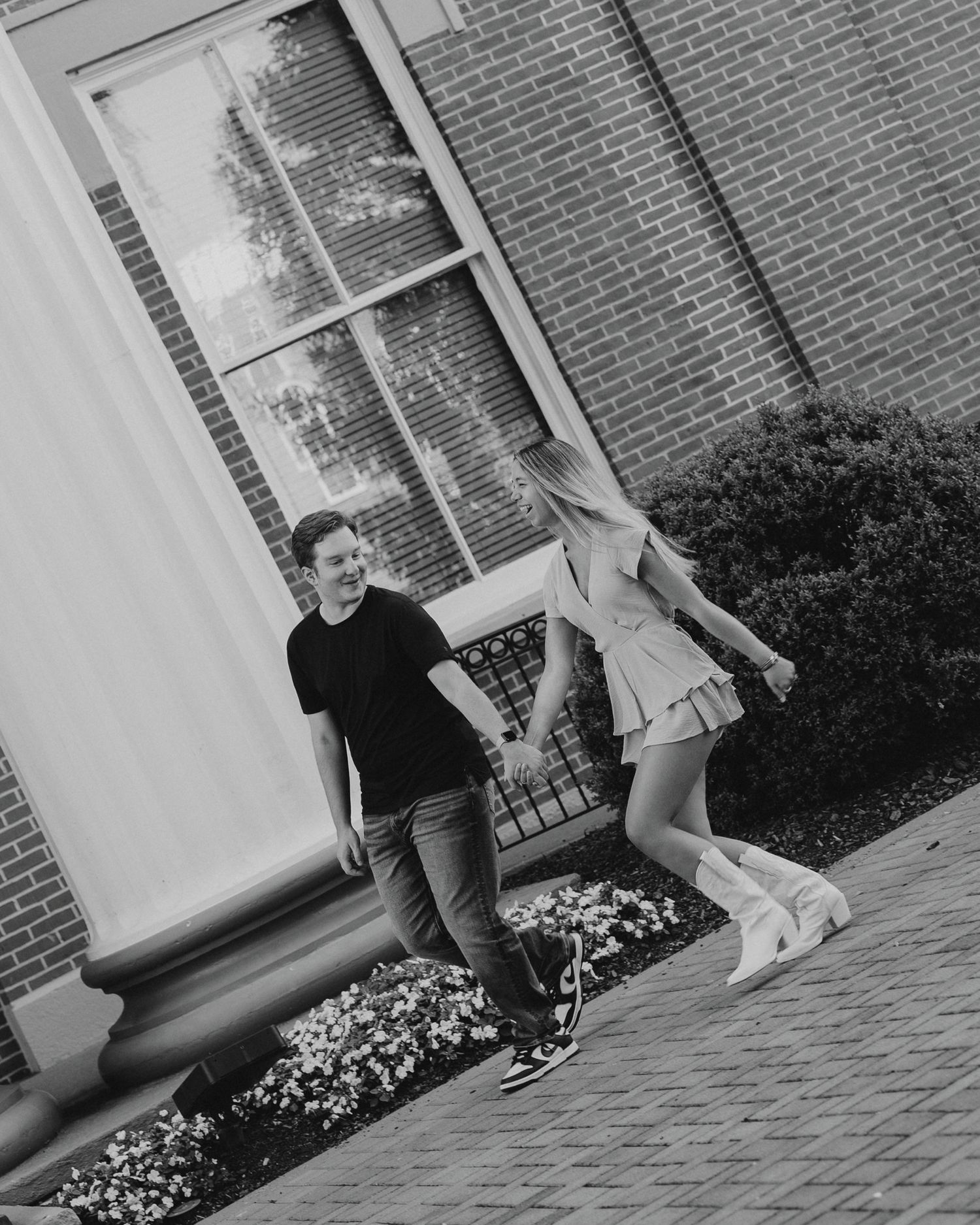 Black and white photo of playful couple holding hands outside brick building.