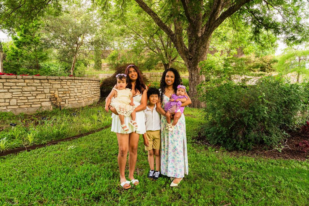 Family of five posing outdoors in lush green garden with large tree, two women holding babies and young boy standing between them.