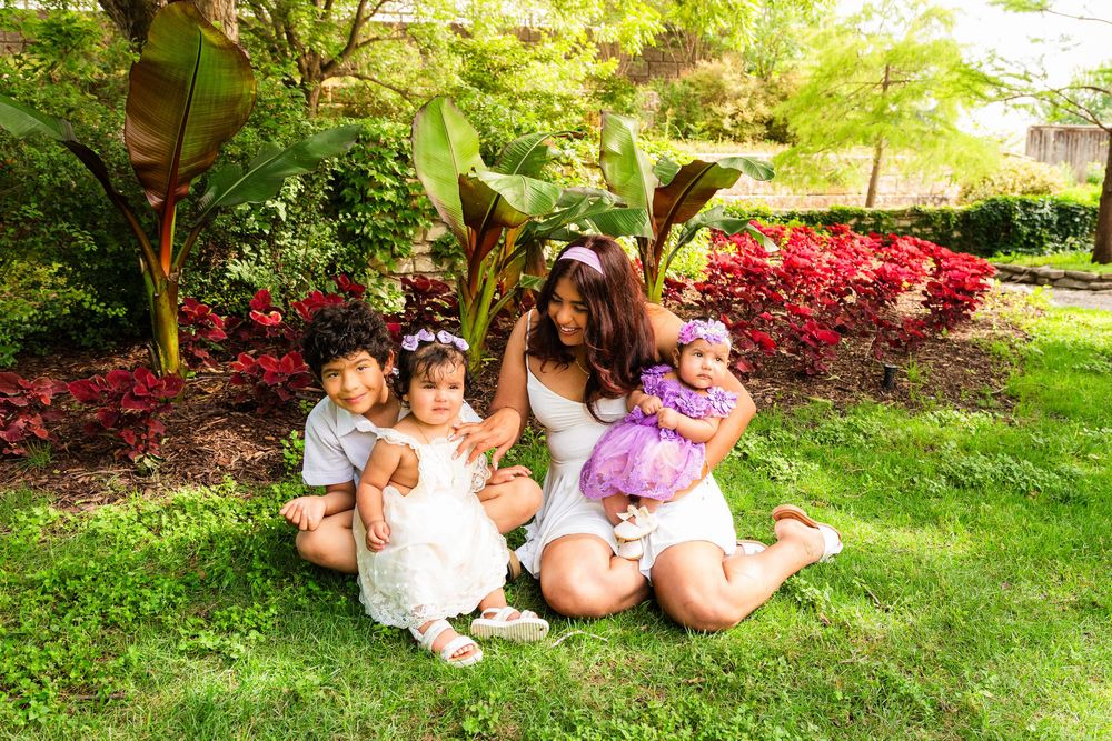A woman sits on green grass with three young children surrounded by colorful garden flowers and lush tropical plants.