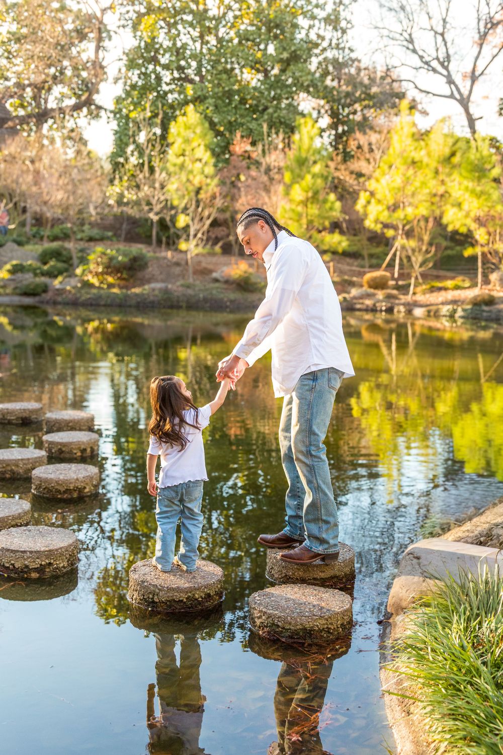 Father helps daughter cross stepping stones over a serene pond in a lush green park on a sunny day.